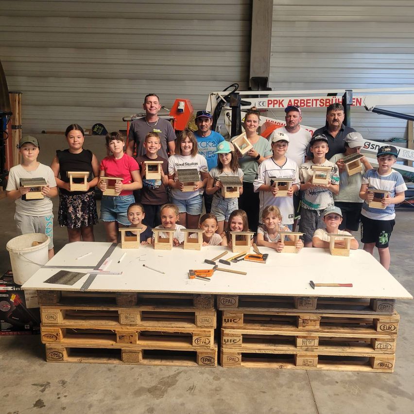Eine Gruppe von Kindern und Erwachsenen steht in einer Werkstatt mit holzernen Vogelhäuschen. Sie lächeln und posieren für ein Foto mit ihren Vogelhäuschen. Ein Eimer und ein Tisch stehen vor ihnen, mit Werkzeugen und anderen Gegenständen darauf.