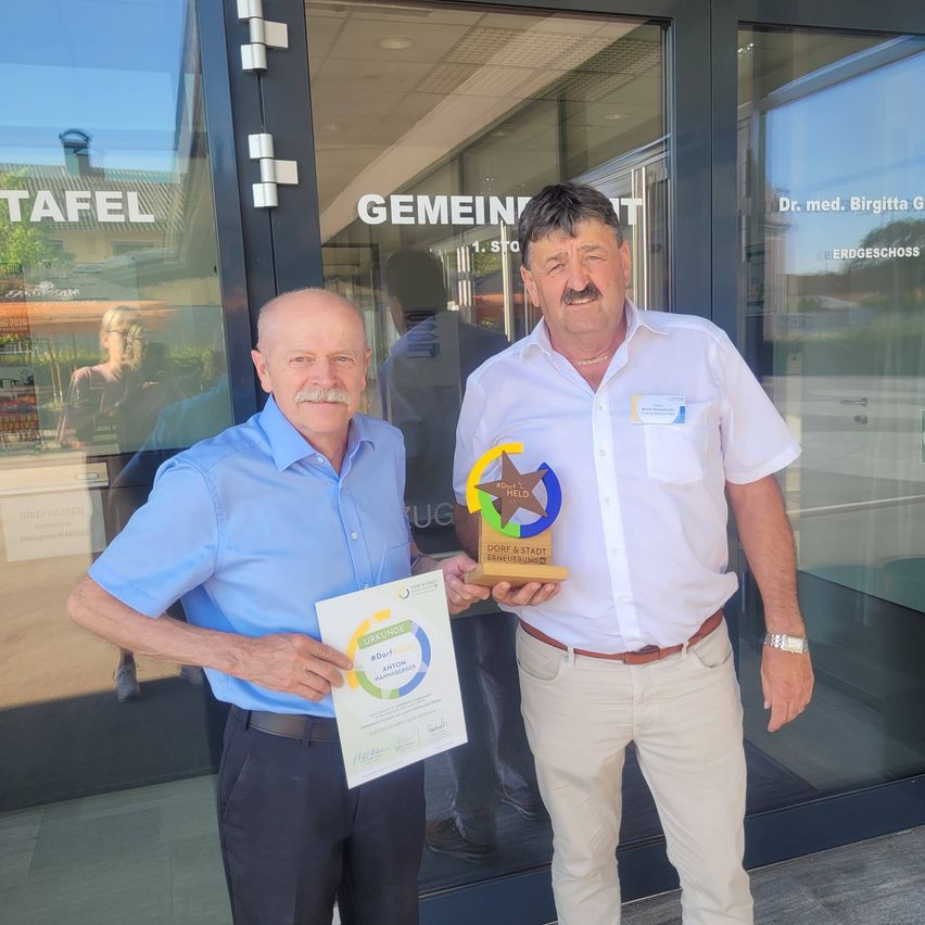 Zwei Männer stehen vor einer Glastür und halten eine Trophäe und ein Zertifikat. Der Mann auf der linken Seite trägt ein blaues Hemd, der andere ein weißes. Ein Gebäude mit Schildern befindet sich hinter ihnen.