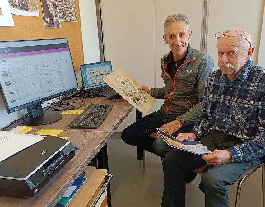 Zwei ältere Männer sitzen vor einem Schreibtisch mit Computer, Tastatur und Drucker. Einer hält ein altes Dokument, während der andere ein Buch liest.