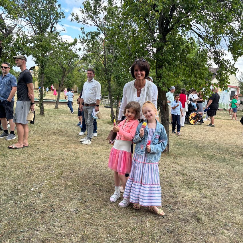 Eine Frau und zwei Mädchen stehen unter einem Baum, lächeln für ein Foto mit Eiscreme. Im Hintergrund gehen Leute umher, einige mit Kindern, und Bäume säumen die Landschaft.