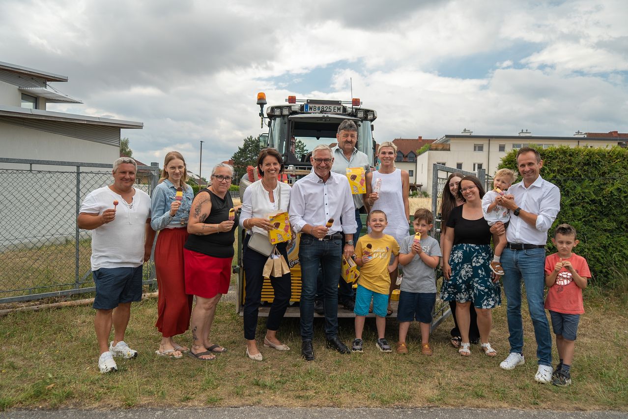 Eine Gruppe von Menschen steht zusammen und hält Eispops in der Hand, mit einem Traktor im Hintergrund. Einige haben Tattoos, und ein Baby wird gehalten. Die Umgebung besteht aus einem Gebäude und einem bewölkten Himmel.