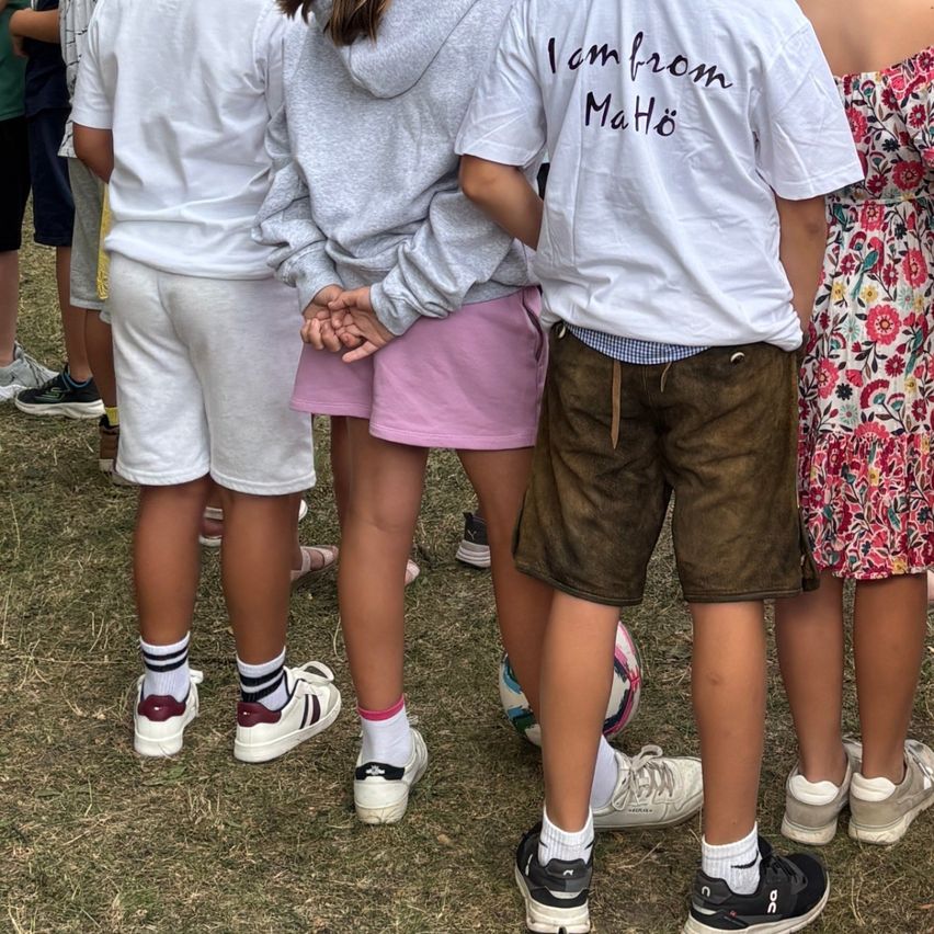 Eine Gruppe von Kindern steht in einem Grasbereich. Ein Kind trägt ein weißes Shirt mit dem Text 'Ich bin aus Mathio'.