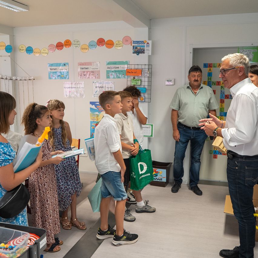 In einem Klassenzimmer versammeln sich eine Gruppe von Kindern und Erwachsenen. Die Kinder halten Bücher und eine grüne Tasche, während ein erwachsener Mann mit ihnen interagiert. Hinter ihnen steht ein Mann in der Nähe einer Tür. Die Wände sind mit bunten Kreisen und Papieren dekoriert.