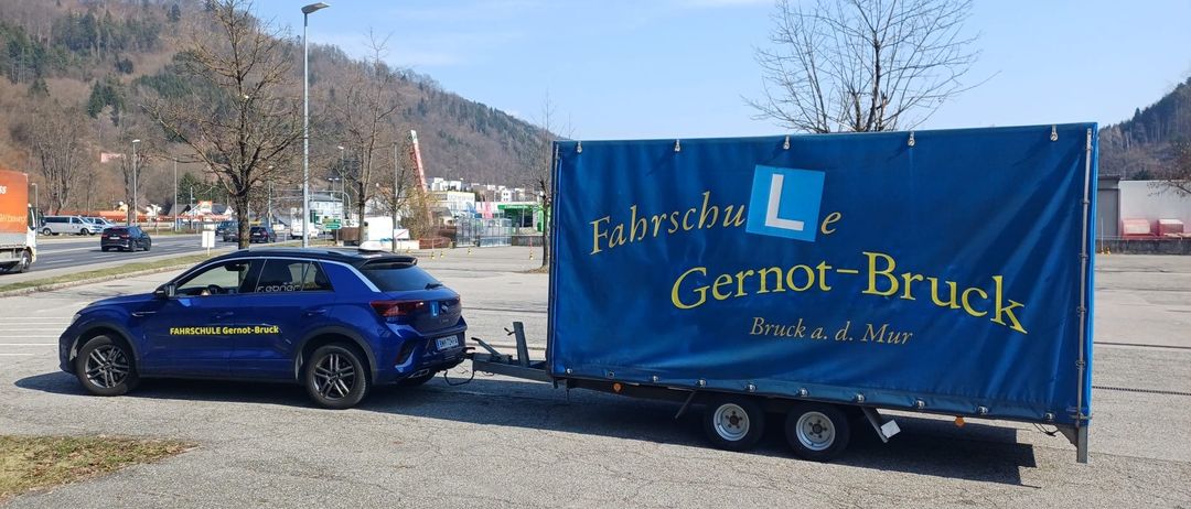 Ein blauer SUV ist neben einem blauen Anhänger mit 'Fahrschule L e Gernot-Brück' darauf geparkt. Im Hintergrund sind Bäume, ein Hügel und eine Straße mit Autos.