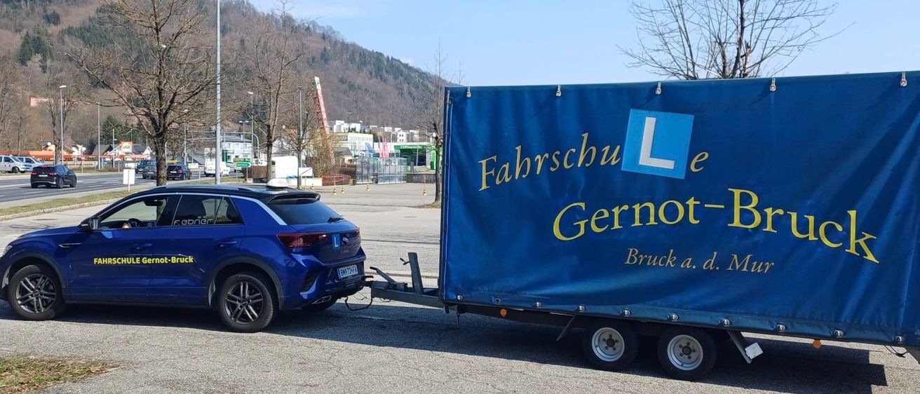 Ein blaues Auto ist in der Nähe eines Anhängers geparkt, der eine blaue Abdeckung mit dem Text 'Fahrschule Gerno Bruck' hat. Das Auto hat ein Nummernschild und einen Aufkleber. Im Hintergrund befinden sich ein Berg, Gebäude und Bäume.