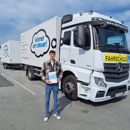 Ein Mann steht vor einem weißen Mercedes-Benz-Lkw mit der Aufschrift 'FAHRSCHULE' auf der Front, hält ein Buch in der Hand. Dahinter stehen zwei weitere Lkw mit Logos. Der Himmel ist bewölkt.