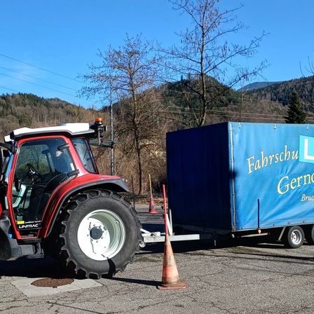 Ein roter Traktor mit einem blauen Anhänger ist auf einer Straße geparkt. Eine Person sitzt im Inneren des Traktors. Ein Verkehrskegel ist neben dem Traktor platziert. Ein blauer Überzug ist auf dem Anhänger, und etwas Text ist darauf geschrieben. Der Berg befindet sich hinter den Bäumen.