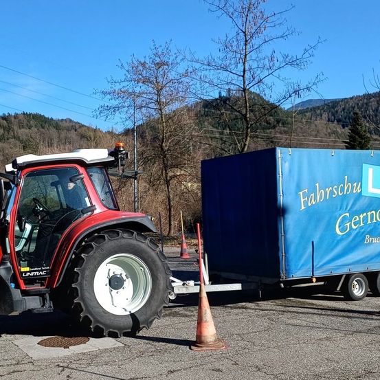 Ein roter Traktor mit einem blauen Anhänger ist auf einer Straße geparkt. Eine Person sitzt im Inneren des Traktors. Ein Verkehrskegel ist neben dem Traktor platziert. Ein blauer Überzug ist auf dem Anhänger, und etwas Text ist darauf geschrieben. Der Berg befindet sich hinter den Bäumen.
