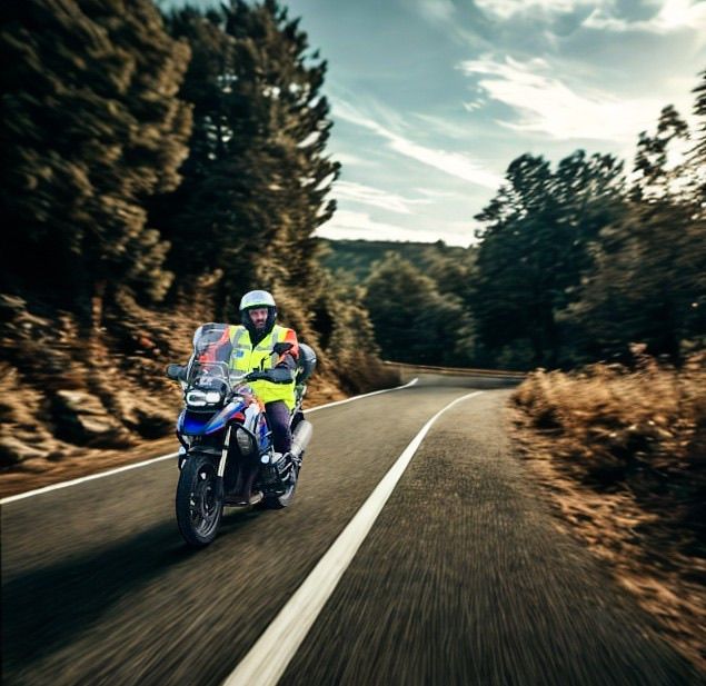 Ein Mann fährt mit einem Motorrad auf einer kurvenreichen Straße und trägt einen Helm und eine gelbe Weste. Die Straße ist von Bäumen und Büschen umgeben.