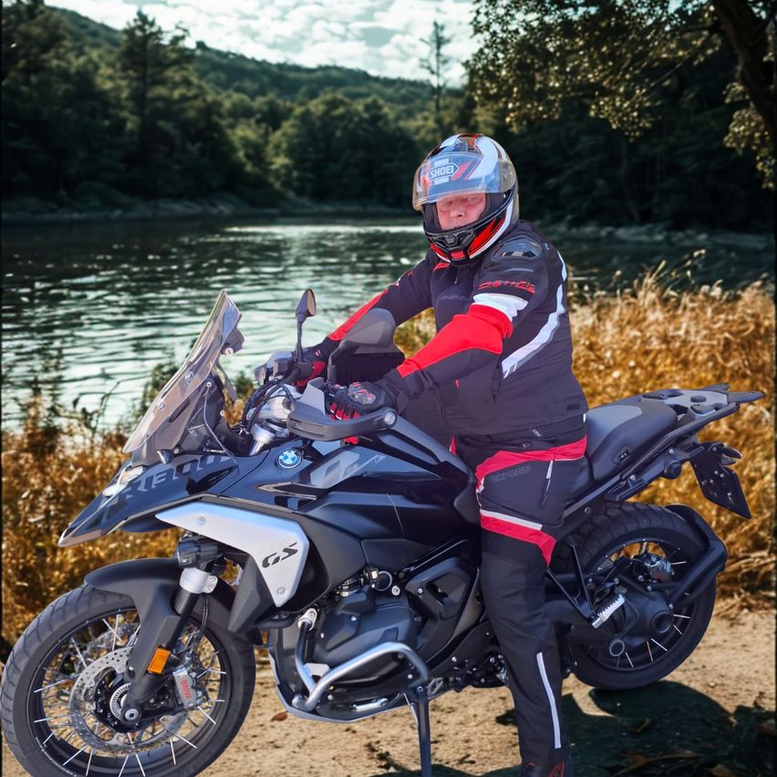 Ein Mann in einem Helm und voller Motorradausrüstung posiert neben einer BMW-Maschine an einem sonnigen Tag am Fluss.