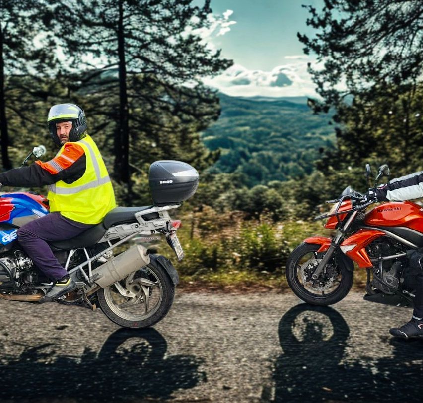 Zwei Motorradfahrer fahren auf einer Straße, einer auf einem blauen Motorrad mit einem Gepäckträger und der andere auf einem roten Motorrad. Sie tragen Helme und Jacken. Bäume und Berge sind im Hintergrund.