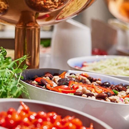 Ein Tisch mit verschiedenen Speisen, darunter eine Schüssel mit Bohnen und Paprika, ein Salat und eine Schüssel mit roten Tomaten. Eine Kupferschüssel befindet sich ebenfalls auf dem Tisch.