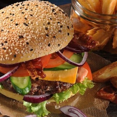 Ein Nahaufnahme eines Hamburgers mit Speck, Tomate, Gurke und Zwiebel auf einem Sesambrötchen, mit Pommes Frites im Hintergrund.