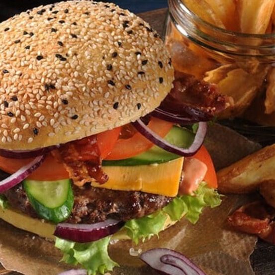 Ein Nahaufnahme eines Hamburgers mit Speck, Tomate, Gurke und Zwiebel auf einem Sesambrötchen, mit Pommes Frites im Hintergrund.