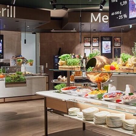 Das Innere eines Restaurants mit einem Buffettisch. Verschiedene Gerichte sind ausgestellt, einschließlich Obst, Gemüse und Salate. Ein Tafel hängt von der Decke mit einem Menü.