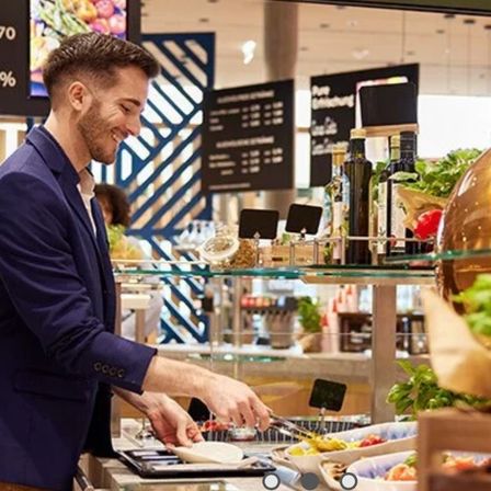 Ein Mann in einem blauen Anzug lächelt und füllt Essen in einem Restaurant. Dahinter befinden sich Flaschen und ein Salatbuffet.