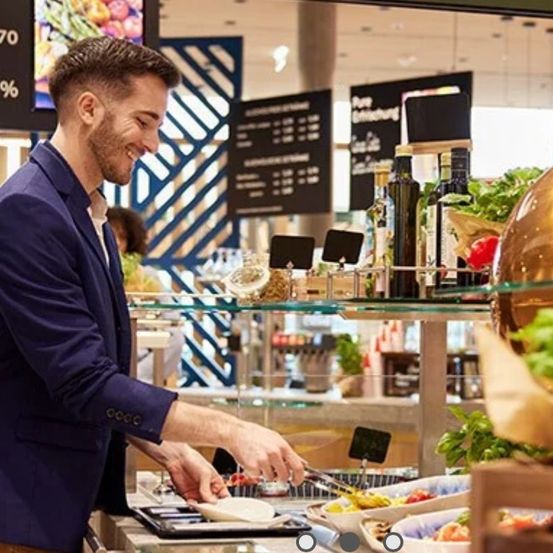 Ein Mann in einem blauen Anzug lächelt und füllt Essen in einem Restaurant. Dahinter befinden sich Flaschen und ein Salatbuffet.