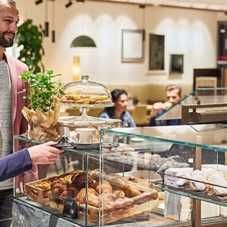 Ein Mann und eine Frau sind in einer Bäckerei und betrachten die ausgestellten Gebäcke auf einem Glastresen. Der Mann hält ein Tablet, während die Frau die Gebäcke betrachtet. Im Hintergrund trinkt ein anderer Mann aus einer Tasse.