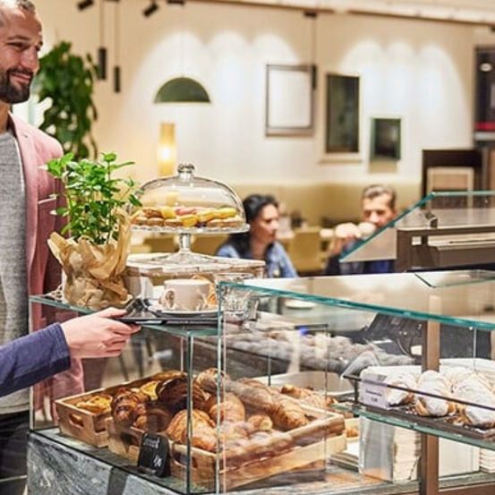 Ein Mann und eine Frau sind in einer Bäckerei und betrachten die ausgestellten Gebäcke auf einem Glastresen. Der Mann hält ein Tablet, während die Frau die Gebäcke betrachtet. Im Hintergrund trinkt ein anderer Mann aus einer Tasse.
