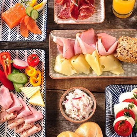 Ein Holztisch zeigt verschiedene Teller mit Essen. Ein Teller hat Lachs, Gurken und Eier. Ein anderer Teller hat Fleisch, Käse und ein Brötchen. Ein dritter Teller hat geschnittenes Fleisch, Käse und Gemüse. Ein vierter Teller hat ein Sandwich mit Tomaten und Käse.