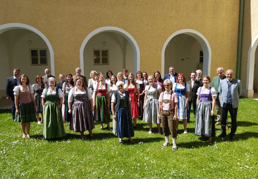 Eine Gruppe von Menschen in traditioneller bayerischer Kleidung posiert für ein Foto vor einem Gebäude mit Bogenfenstern und -türen.