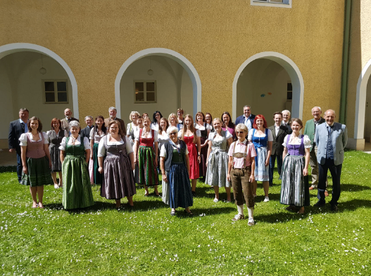 Eine Gruppe von Menschen in traditioneller bayerischer Kleidung posiert für ein Foto vor einem Gebäude mit Bogenfenstern und -türen.