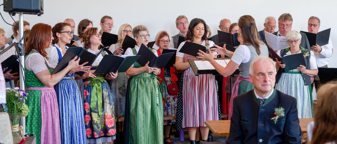 Ein Chor aus Frauen und Männern in traditioneller Kleidung singt zusammen mit Notenblättern in einem großen Raum.