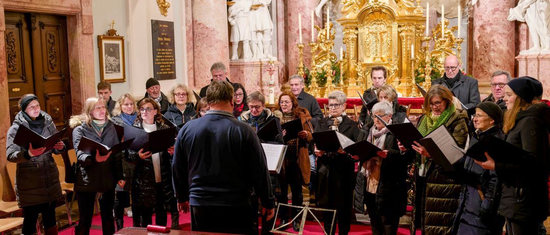 Ein Chor von Menschen steht auf einem roten Teppich vor einem Klavier und singt, während sie in die Notenblätter schauen. Dahinter befindet sich ein goldener Altar und weiße Statuen.