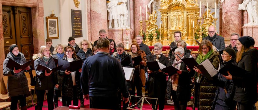 Ein Chor von Menschen steht auf einem roten Teppich vor einem Klavier und singt, während sie in die Notenblätter schauen. Dahinter befindet sich ein goldener Altar und weiße Statuen.