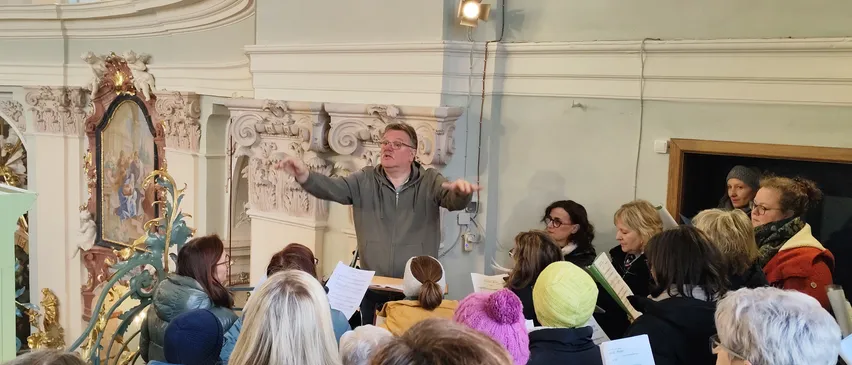 Ein Mann mit Brille dirigiert einen Chor in einem Saal. Er gestikuliert mit den Händen, während mehrere Menschen vor ihm sitzen. Einige tragen Mützen. Eine Lampe ist an der Wand hinter ihm montiert.