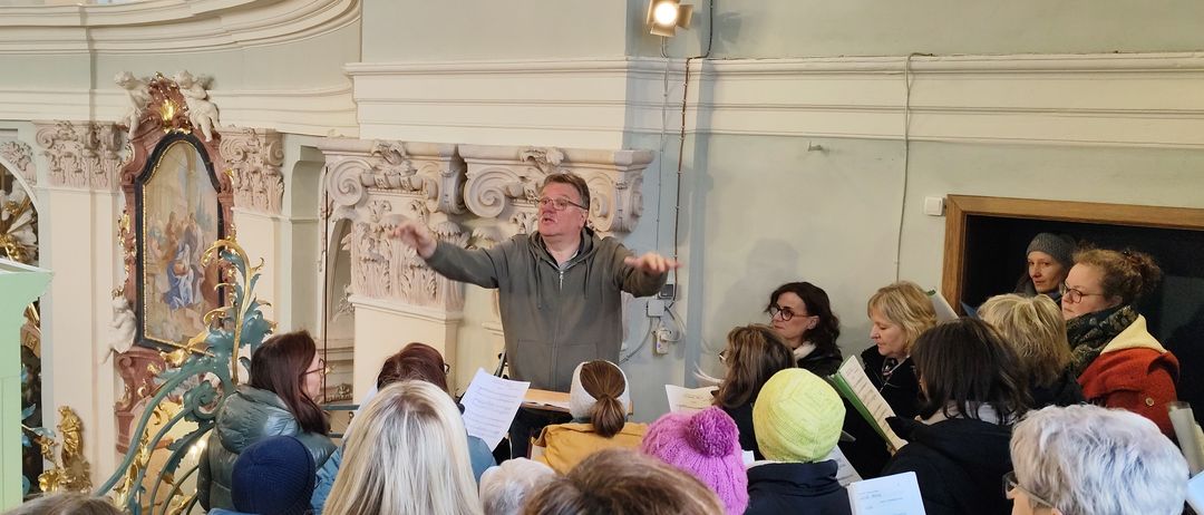 Ein Mann mit Brille dirigiert einen Chor in einem Saal. Er gestikuliert mit den Händen, während mehrere Menschen vor ihm sitzen. Einige tragen Mützen. Eine Lampe ist an der Wand hinter ihm montiert.