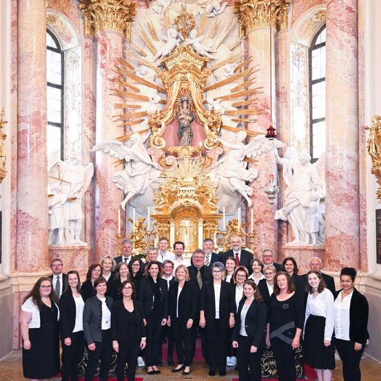 Eine Gruppe von Menschen, in formeller Kleidung, steht vor einem Kirchenaltar. Der Altar ist mit einer goldenen Struktur und Statuen geschmückt. Der Raum hat rosa Marmorwände und Fenster.