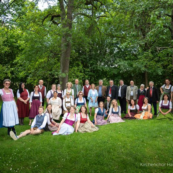 Eine Gruppe von Menschen, einige in traditioneller Kleidung, posiert für ein Foto im Park. Viele tragen Sonnenbrillen, und einige tragen Krawatten. Bäume stehen hinter ihnen.