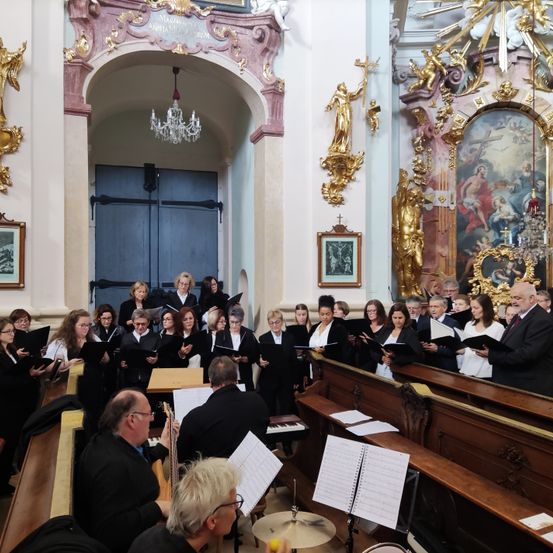 Ein Chor singt in einer Kirche. Viele Menschen halten Bücher und singen. Die Kirche hat goldene Statuen und Gemälde.