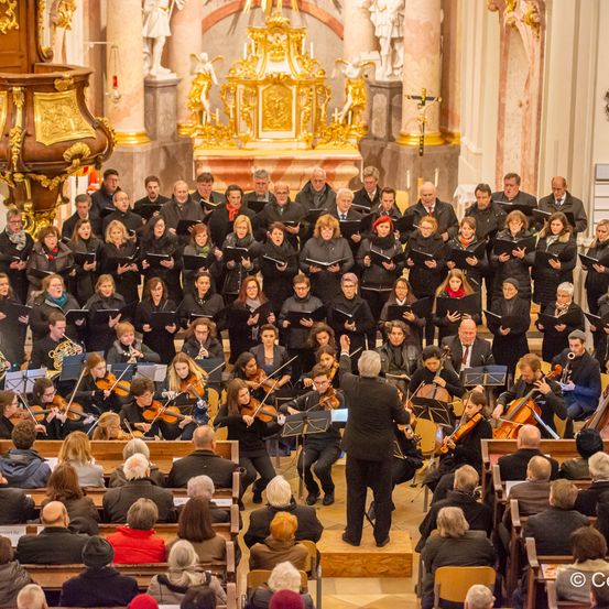Ein Chor tritt in einer Kirche auf, angeführt von einem Dirigenten. Der Chor steht vor einem Altar mit goldenen Dekorationen. Das Publikum sitzt auf Bänken vor dem Chor.