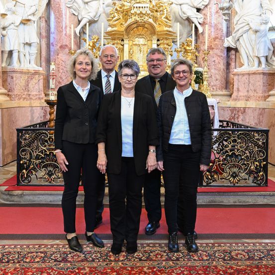 Fünf Personen stehen vor einem kunstvollen Altar in einer Kirche und posieren für ein Foto. Dahinter sind Statuen und Kerzen zu sehen.