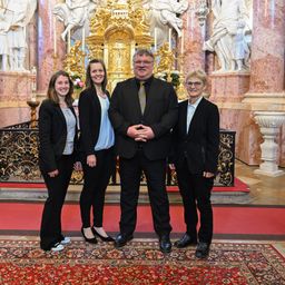 Vier Menschen stehen vor einem Altar in einer Kirche. Sie sind in formeller Kleidung gekleidet. Ein roter Teppich liegt auf dem Boden. Hinter ihnen schmücken Statuen die Wände.