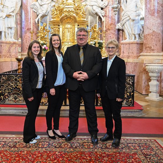 Vier Menschen stehen vor einem Altar in einer Kirche. Sie sind in formeller Kleidung gekleidet. Ein roter Teppich liegt auf dem Boden. Hinter ihnen schmücken Statuen die Wände.