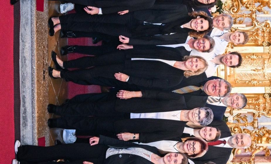 Ein Chor von Personen in Schwarz gekleidet, in zwei Reihen angeordnet, lächelt und posiert für ein Foto in einem großen, prunkvollen Raum.