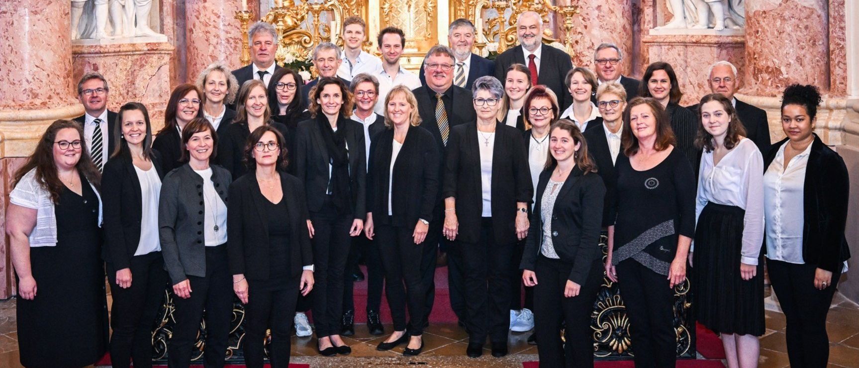 Eine Gruppe formell gekleideter Personen, darunter Männer und Frauen, posiert für ein Foto in einer Kirche. Sie stehen vor einem prächtigen Altar mit goldenem Hintergrund.