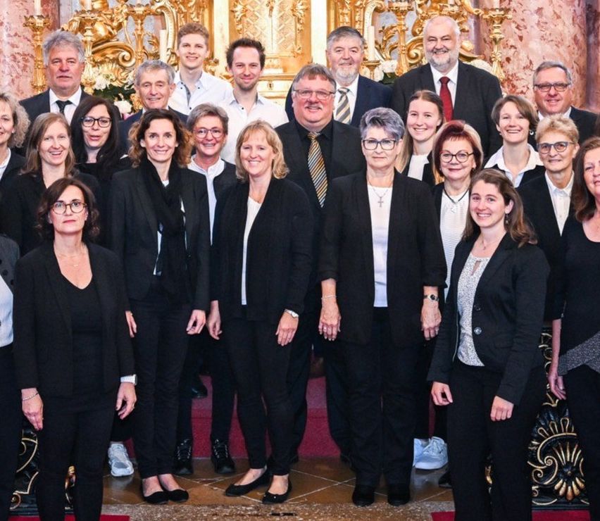 Eine Gruppe formell gekleideter Personen, darunter Männer und Frauen, posiert für ein Foto in einer Kirche. Sie stehen vor einem prächtigen Altar mit goldenem Hintergrund.