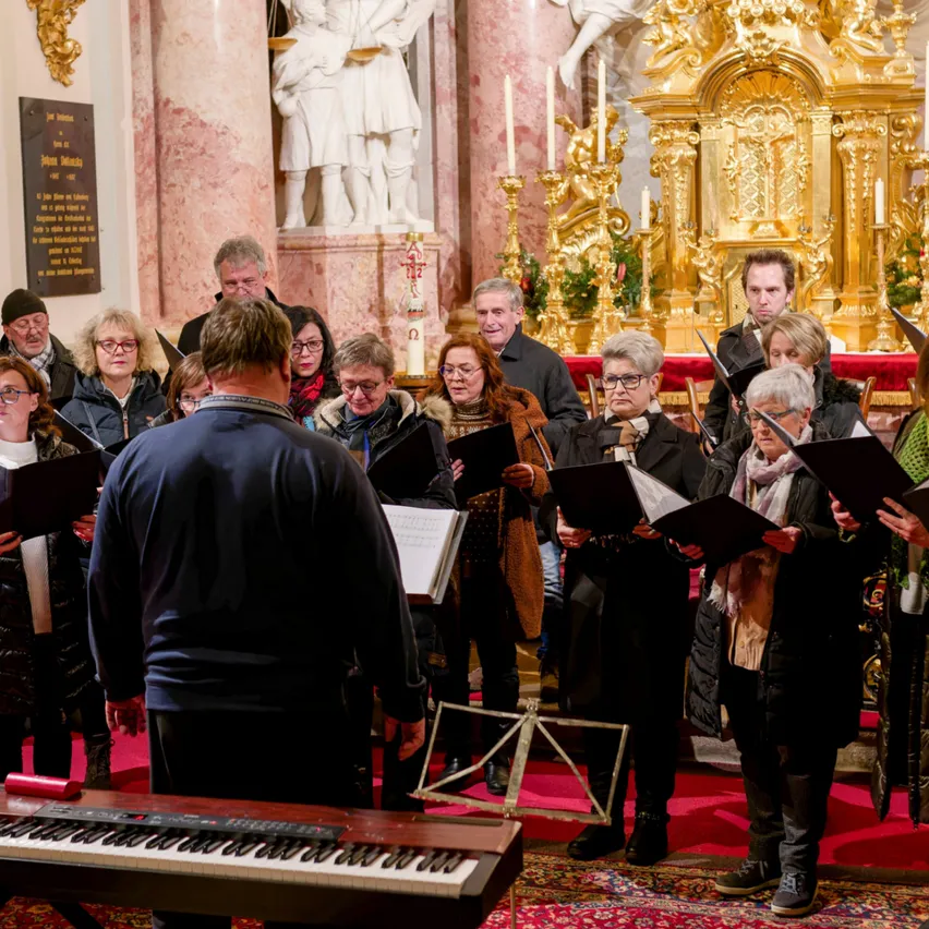 Ein Chor steht in einer Kirche und singt, während er Notenblätter betrachtet. Hinter ihnen befindet sich ein goldener Altar mit Kerzen. Die Kirche ist mit Dekorationen geschmückt.