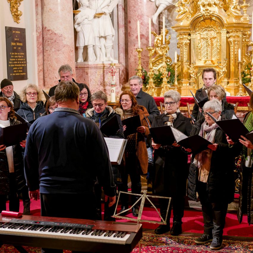 Ein Chor steht in einer Kirche und singt, während er Notenblätter betrachtet. Hinter ihnen befindet sich ein goldener Altar mit Kerzen. Die Kirche ist mit Dekorationen geschmückt.