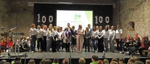 Eine Gruppe von Menschen in weißen Hemden und grünen T-Shirts steht mit Luftballons auf der Bühne, während eine Frau ein Mikrofon hält. Hinter ihnen zeigt eine Projektionsleinwand Text. Kinder sitzen davor.