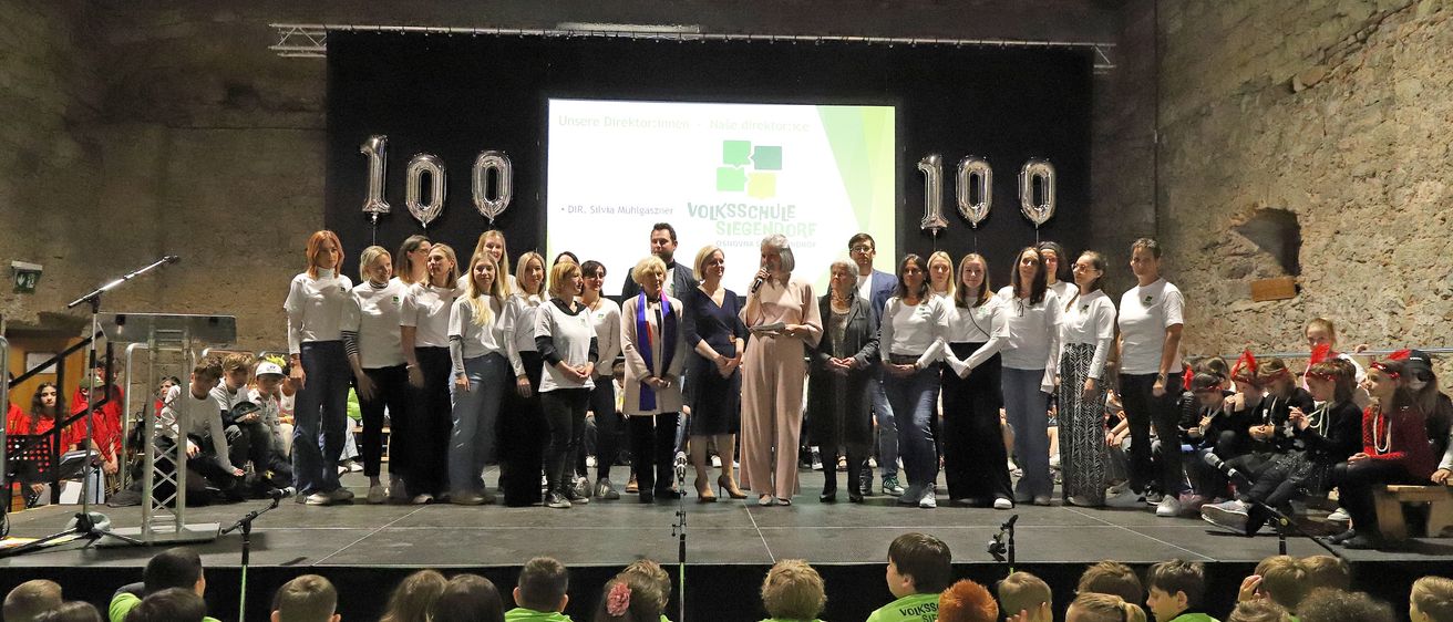 Eine Gruppe von Menschen in weißen Hemden und grünen T-Shirts steht mit Luftballons auf der Bühne, während eine Frau ein Mikrofon hält. Hinter ihnen zeigt eine Projektionsleinwand Text. Kinder sitzen davor.