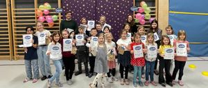 Eine Gruppe von Kindern und Erwachsenen mit Urkunden steht in einem Raum mit Luftballons und einer dekorierten Wand. Einige tragen Turnschuhe und haben Taschen bei sich.