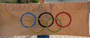 Drei Jungen in grünen Shirts halten ein Banner mit olympischen Ringen und dem Text '1. Olympiade vs. Siegendorf'. Sie stehen vor einem Gebäude und einem Baum.