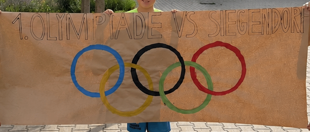 Drei Jungen in grünen Shirts halten ein Banner mit olympischen Ringen und dem Text '1. Olympiade vs. Siegendorf'. Sie stehen vor einem Gebäude und einem Baum.