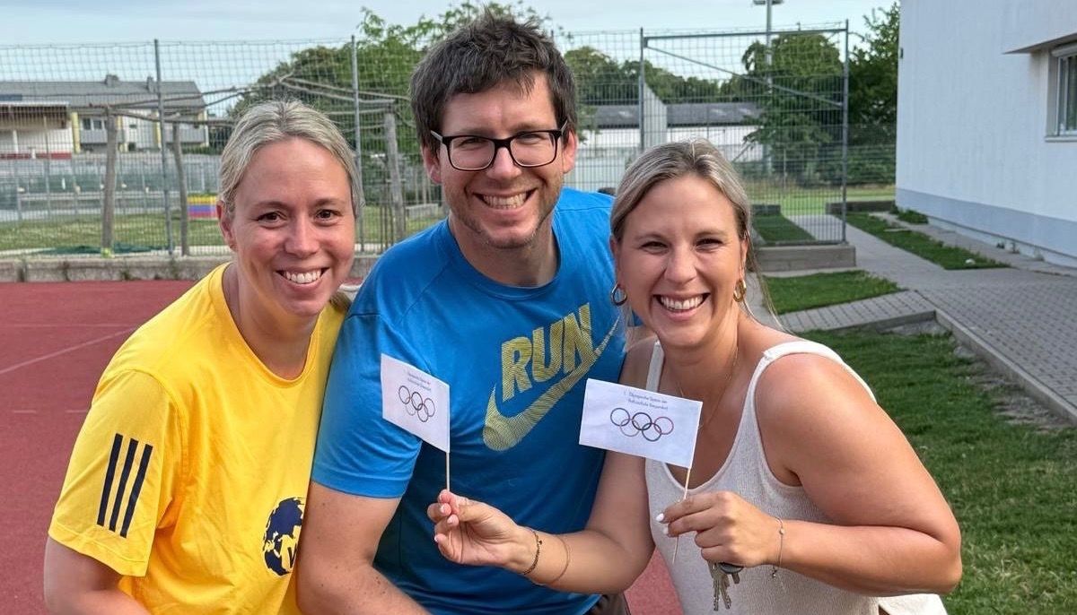 Drei Personen stehen lächelnd in einem Sportbereich. Einer hält olympische Flaggen. Ein Mann trägt ein blaues T-Shirt mit Nike-Logo, während zwei Frauen ein gelbes und ein weißes T-Shirt tragen. Dahinter befindet sich ein Zaun, ein Gebäude und Bäume.