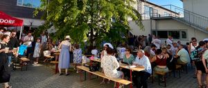 Eine Gruppe von Menschen sitzt unter einem Baum um Tische herum, mit Gebäuden im Hintergrund und einem roten Zelt auf der linken Seite. Sie tragen Freizeitkleidung und einige tragen Hüte.